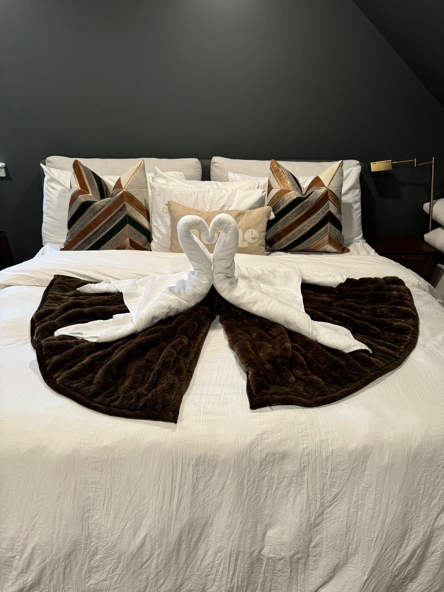 Bed made with white linens and two towel swans forming a heart on a dark throw
