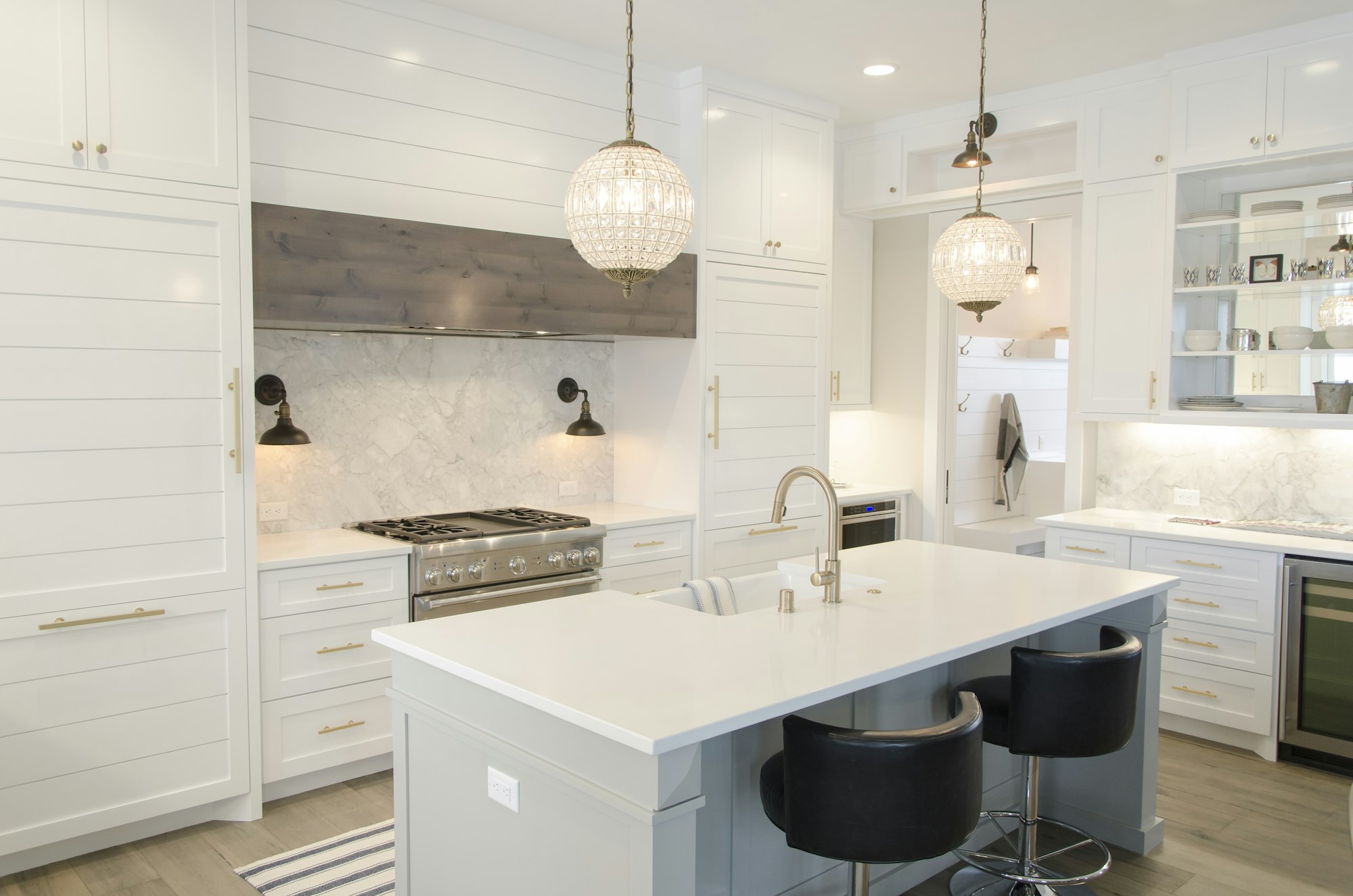A bright white kitchen with a marble island, black stools and glass pendant lights